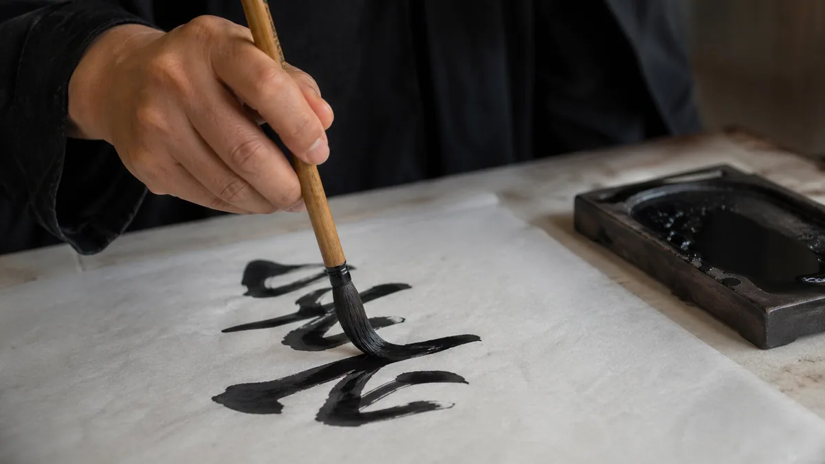 East Asian calligrapher's hand holding a bamboo-handled brush vertically, painting a fluid black ink stroke on rice paper