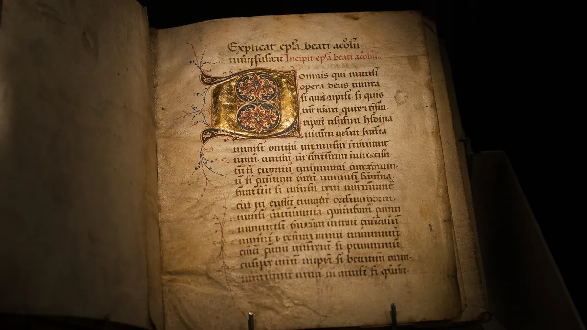 Museum-lit ancient illuminated medieval manuscript page on aged vellum with a gold leaf decorative initial