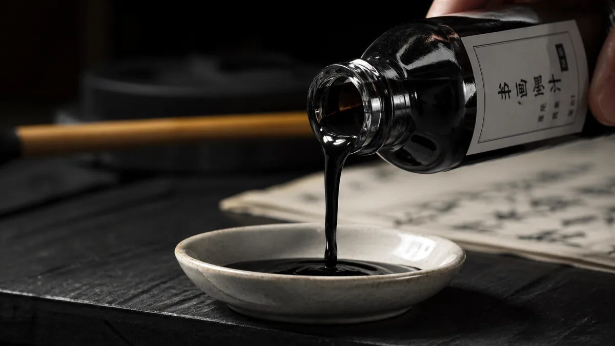 Action shot of black calligraphy ink being poured slowly from a glass bottle into a small ceramic dinky dip dish