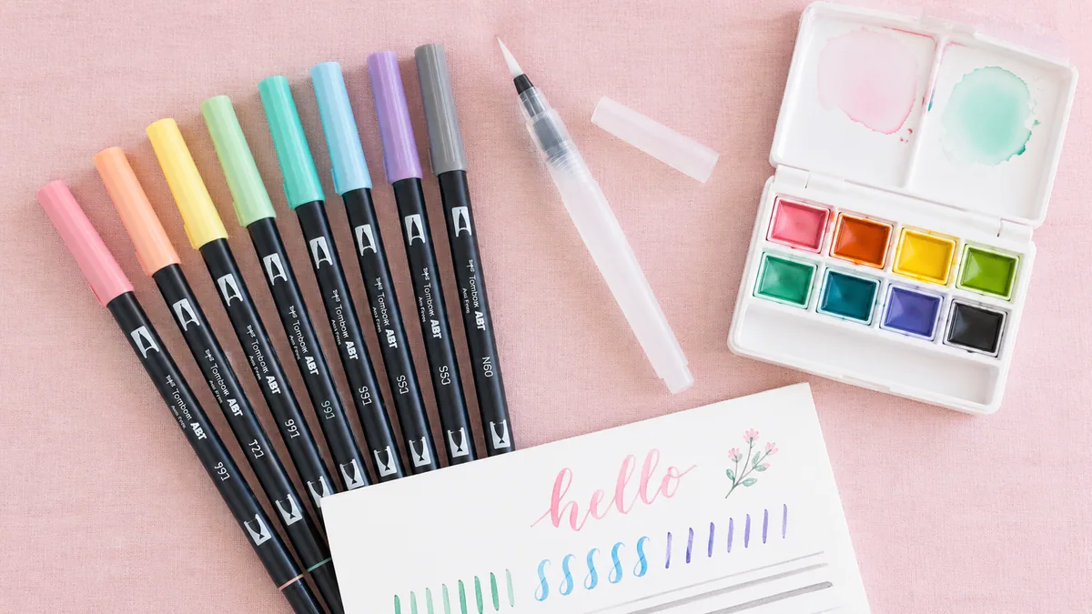 Top-down flat-lay of pastel Tombow brush pens, water brush, and watercolor palette on a soft pink linen background